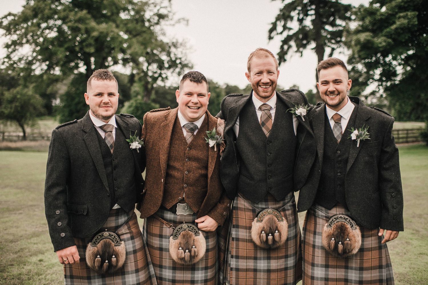 Four groomsmen in matching kilts and formal jackets pose for a photo outdoors.