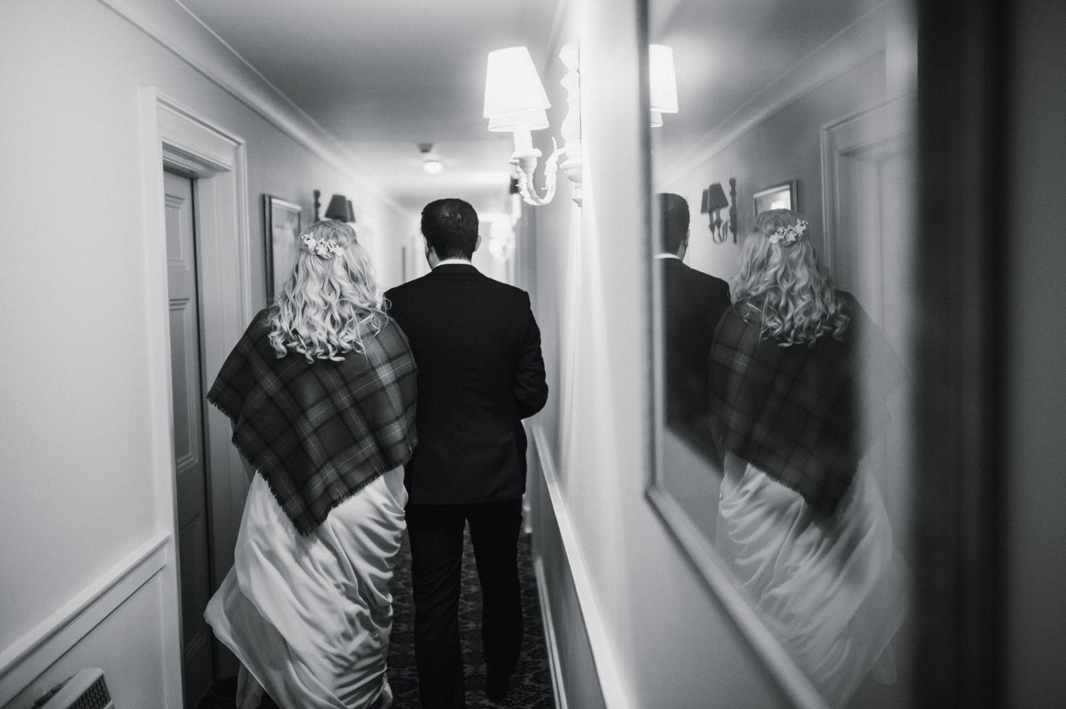 Black and white image of two people walking down hallway with mirror reflections.