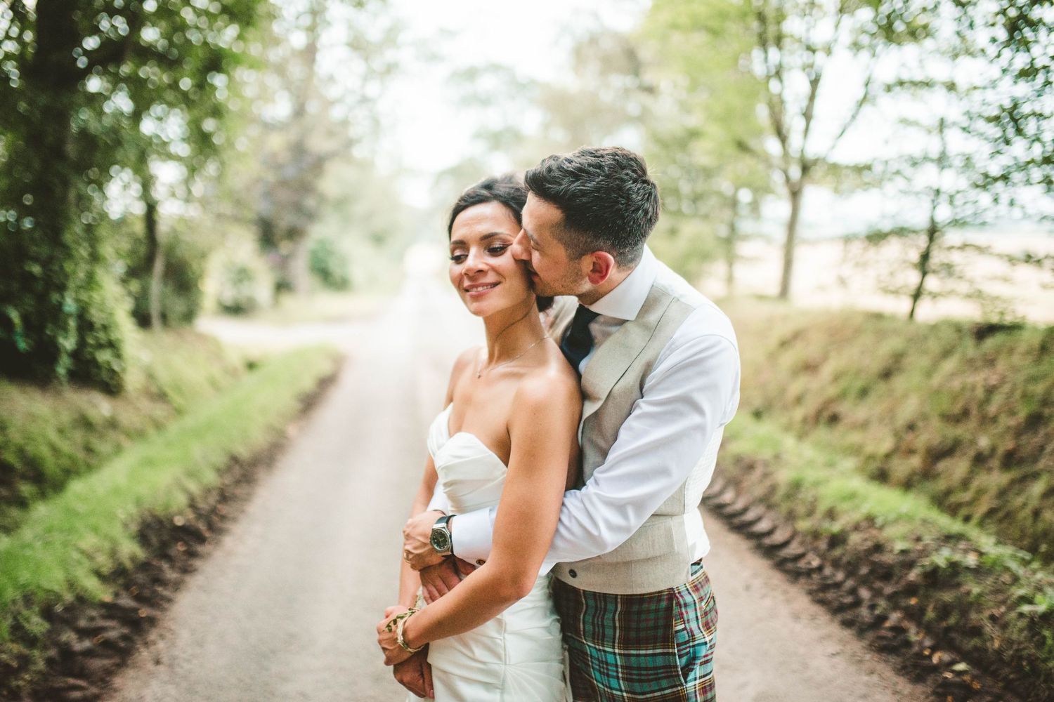 A romantic moment captured between newlyweds on a country lane with the groom wearing traditional Scottish attire.