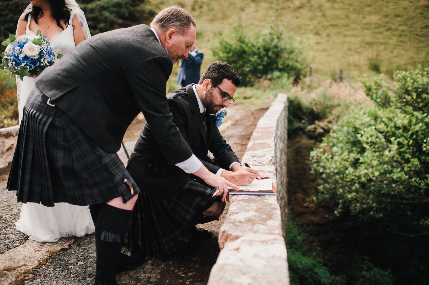 Wedding party members lean over a stone bridge wall during a Scottish countryside ceremony.