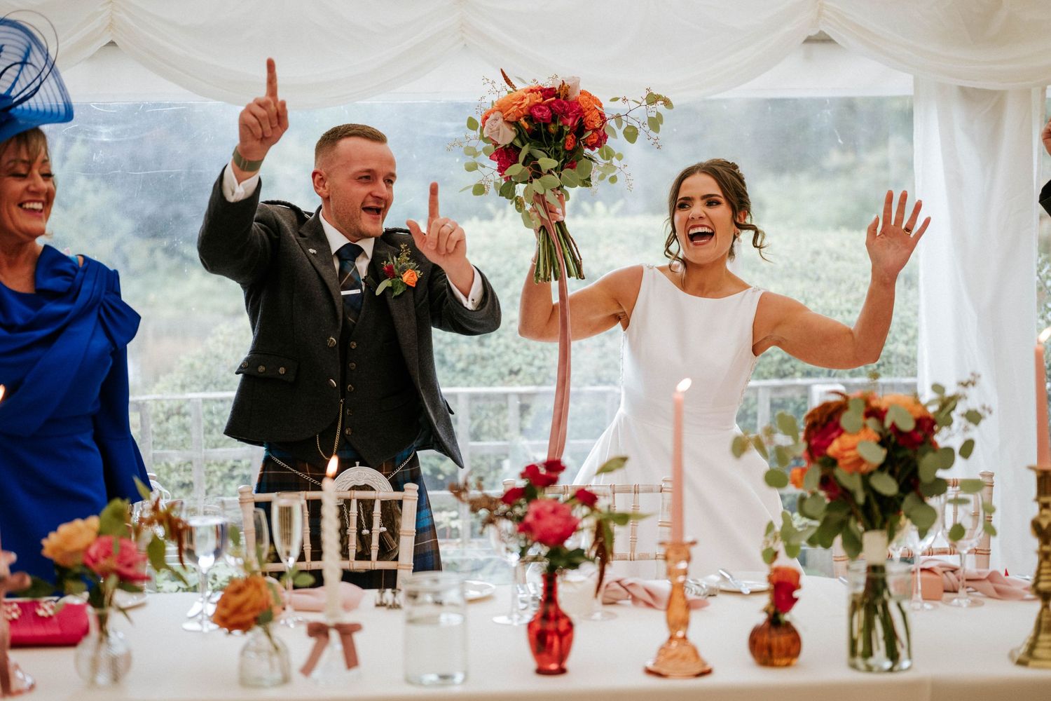 Wedding celebration under a white tent with floral centerpieces and guests showing excitement.