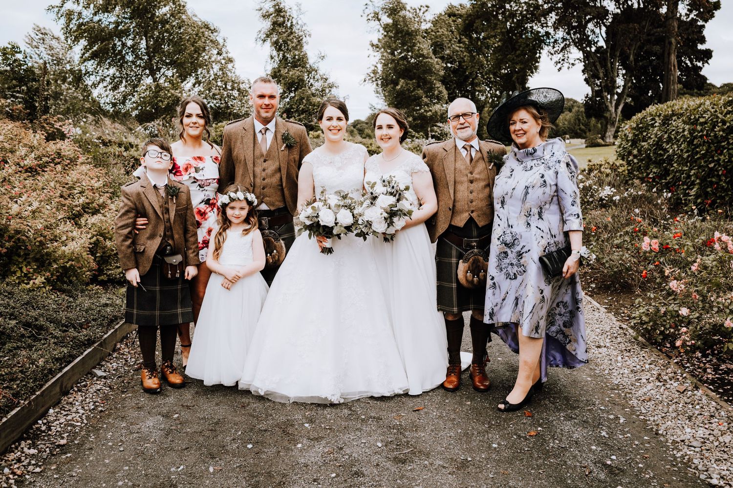 Wedding party poses on a gravel path with mix of formal wear and traditional Scottish attire.