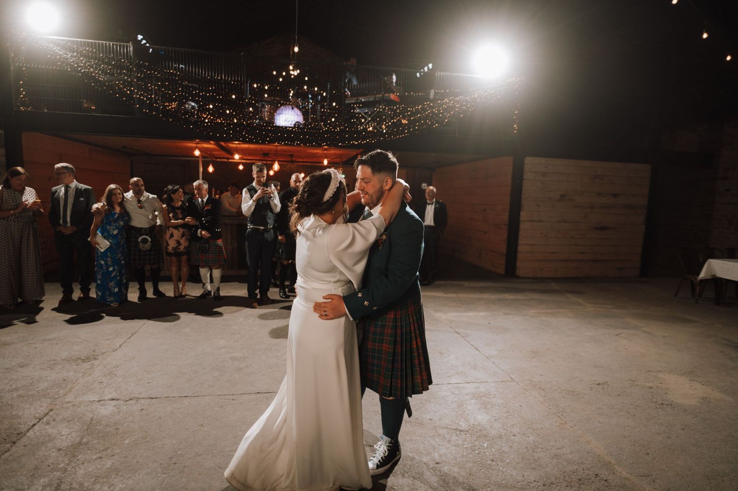 A couple embraces during their wedding dance while guests look on in a rustic venue with overhead lighting.