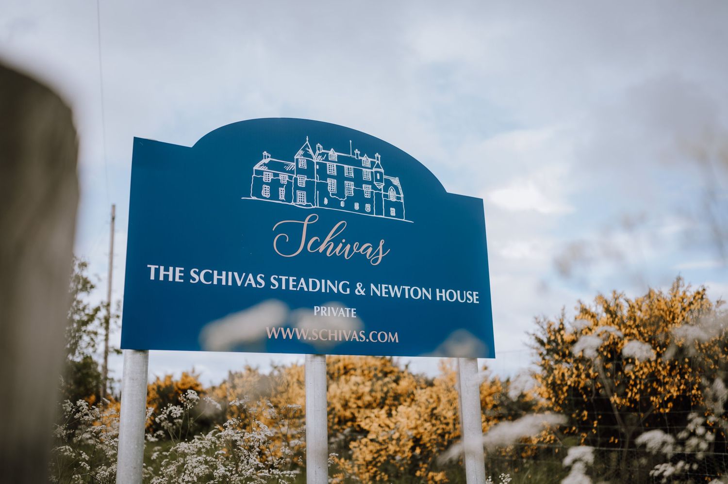 A blue entrance sign for The Schivas Reading & Newton House against autumn foliage.
