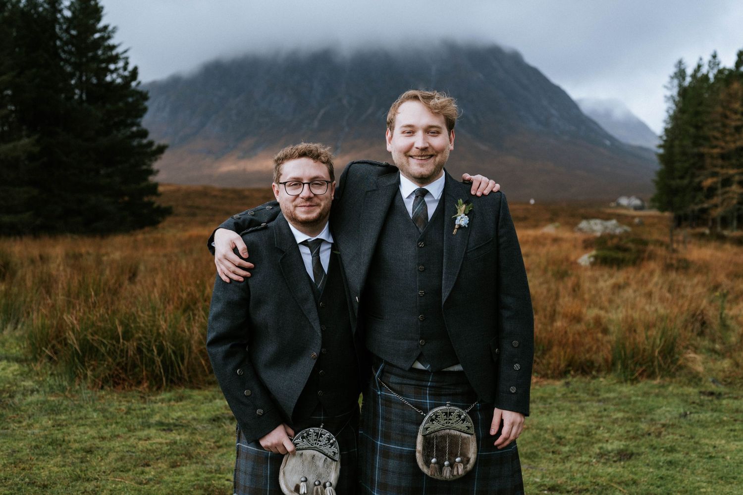 Two people in traditional Scottish kilts and formal wear pose together against mountain backdrop.