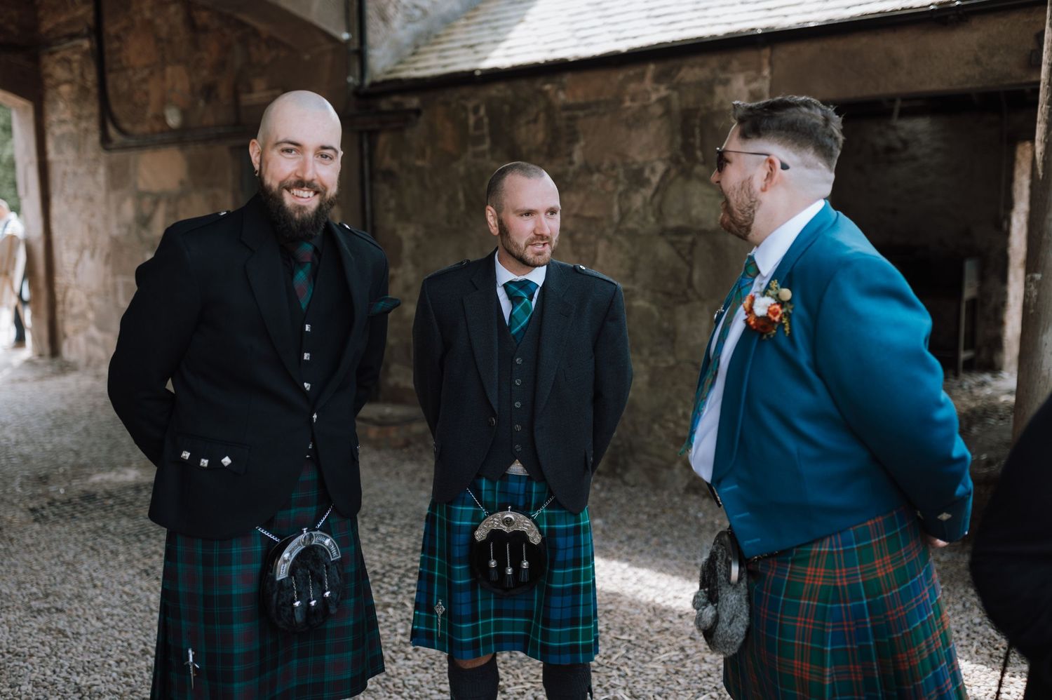 Three men in traditional Scottish kilts and formal attire chat at outdoor event.
