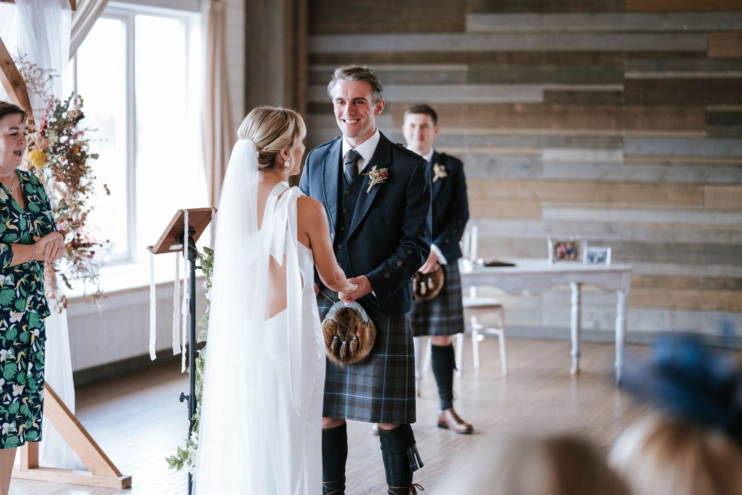 A first look wedding moment unfolds in a bright indoor venue with wooden accents and floral decorations.