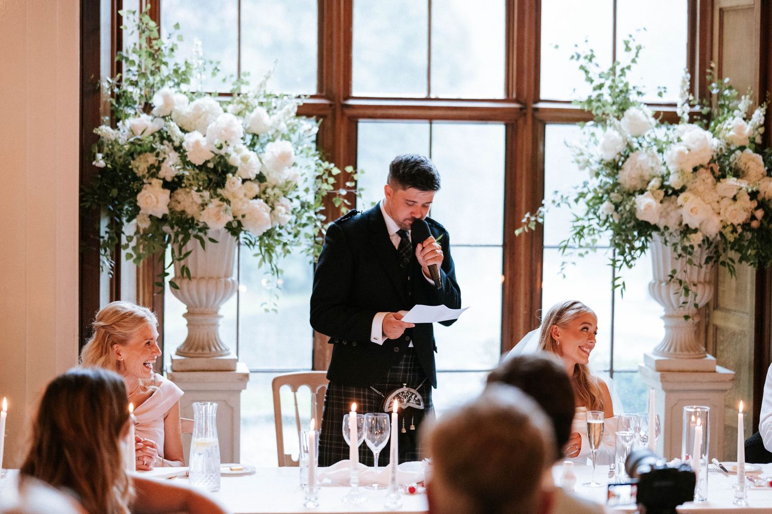 A speaker delivers a speech at an elegant wedding reception decorated with tall white floral arrangements.
