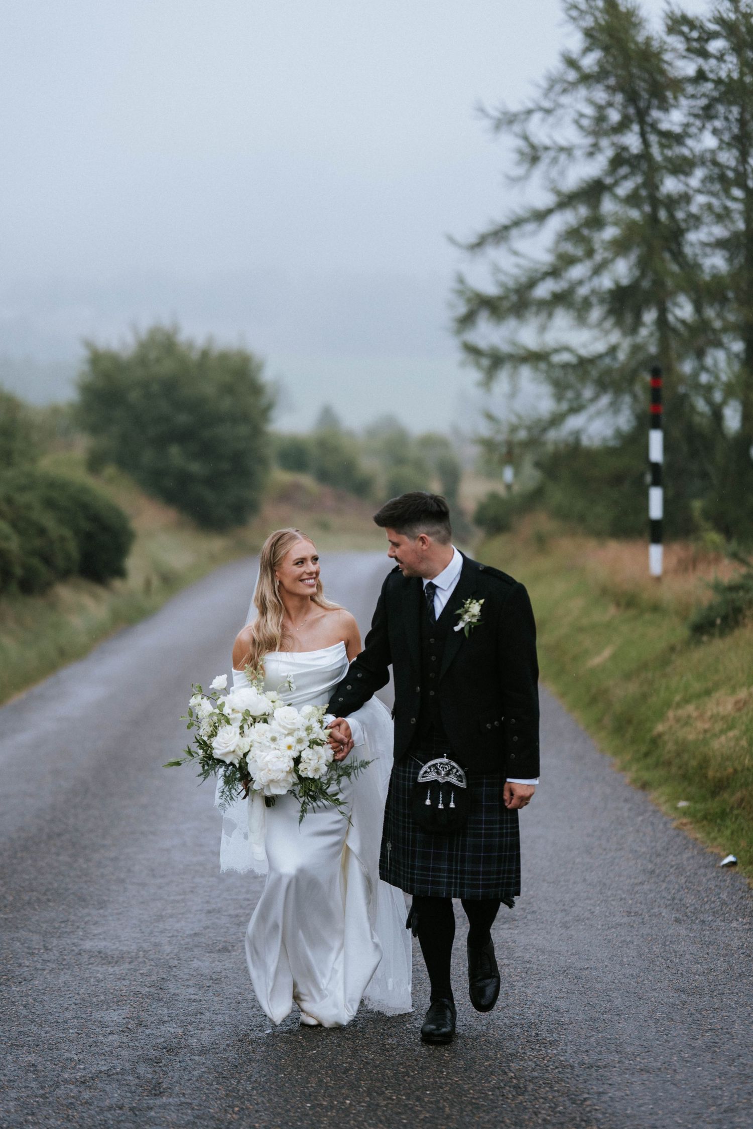 A bride and groom share a joyful moment while walking down a scenic rural path in Scotland.