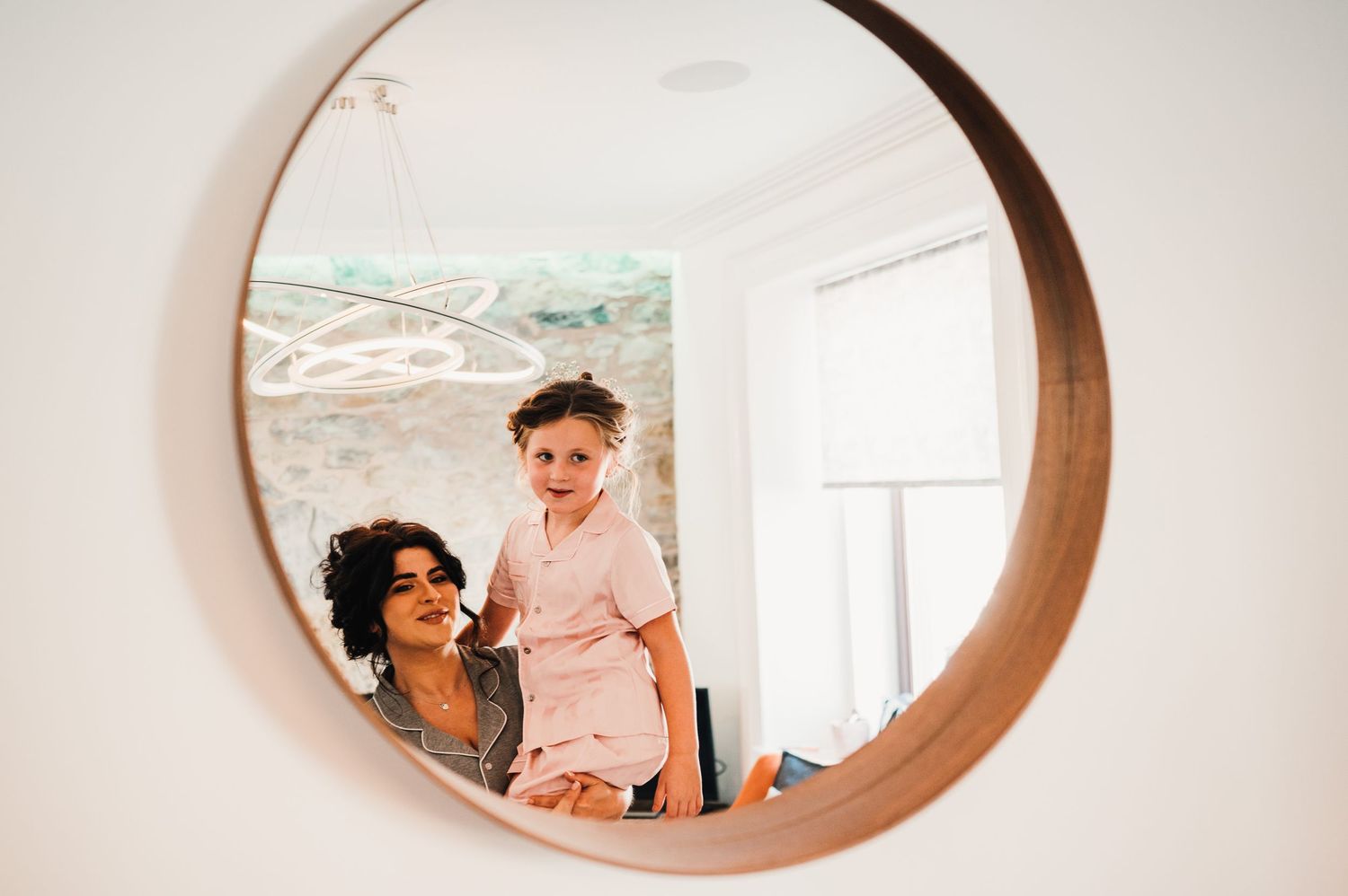 A reflection in a round wooden mirror shows two people laughing together in a bright interior space.