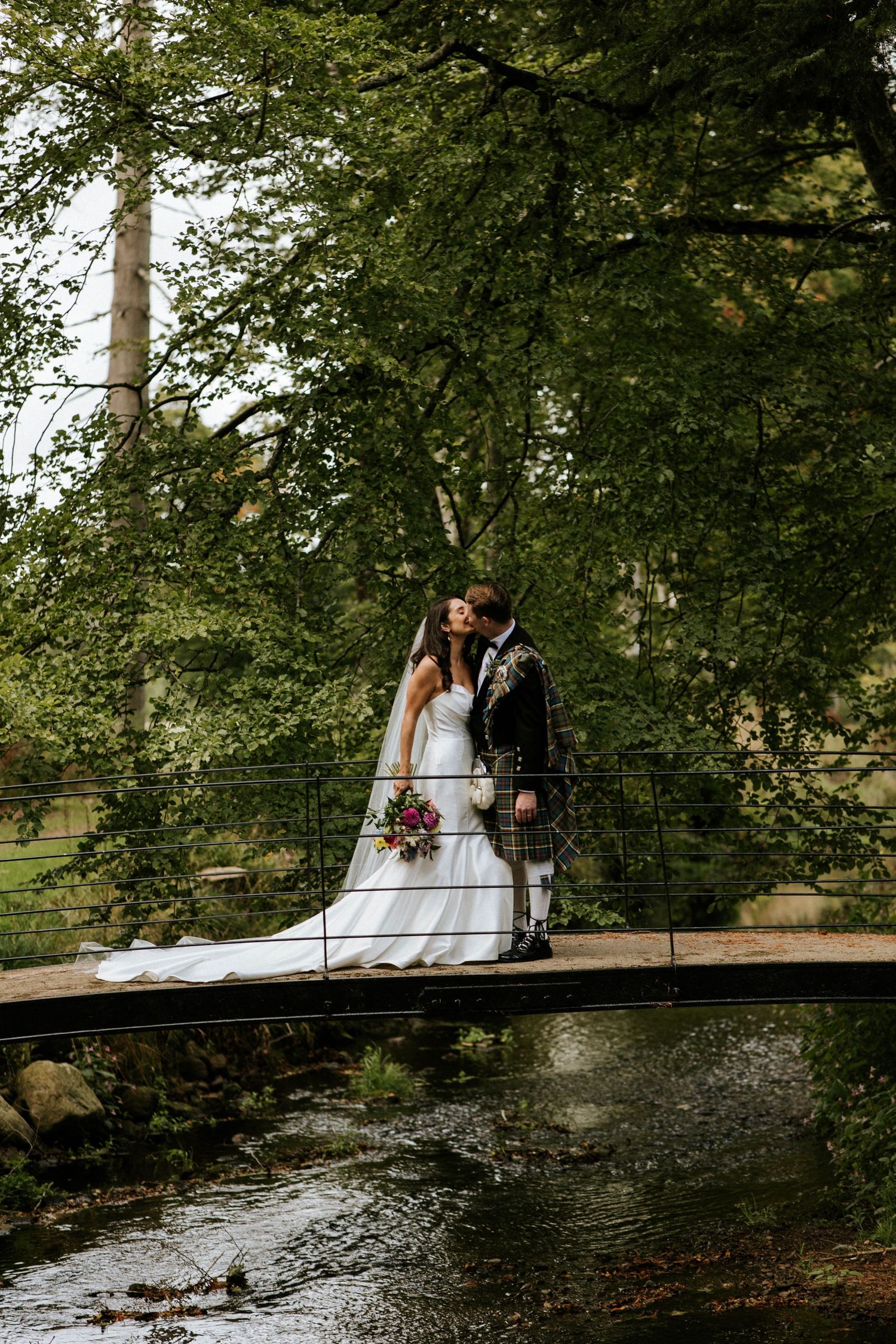 A couple sits on a rustic wooden bridge over a pond in a lush wooded setting during their wedding day.