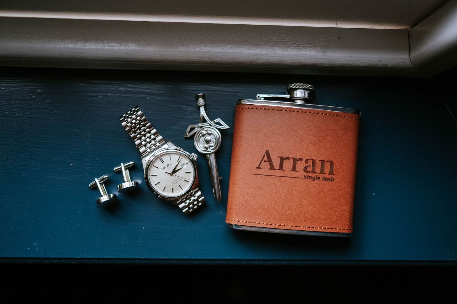 Wedding accessories including cufflinks, flask and watch arranged on dark surface.