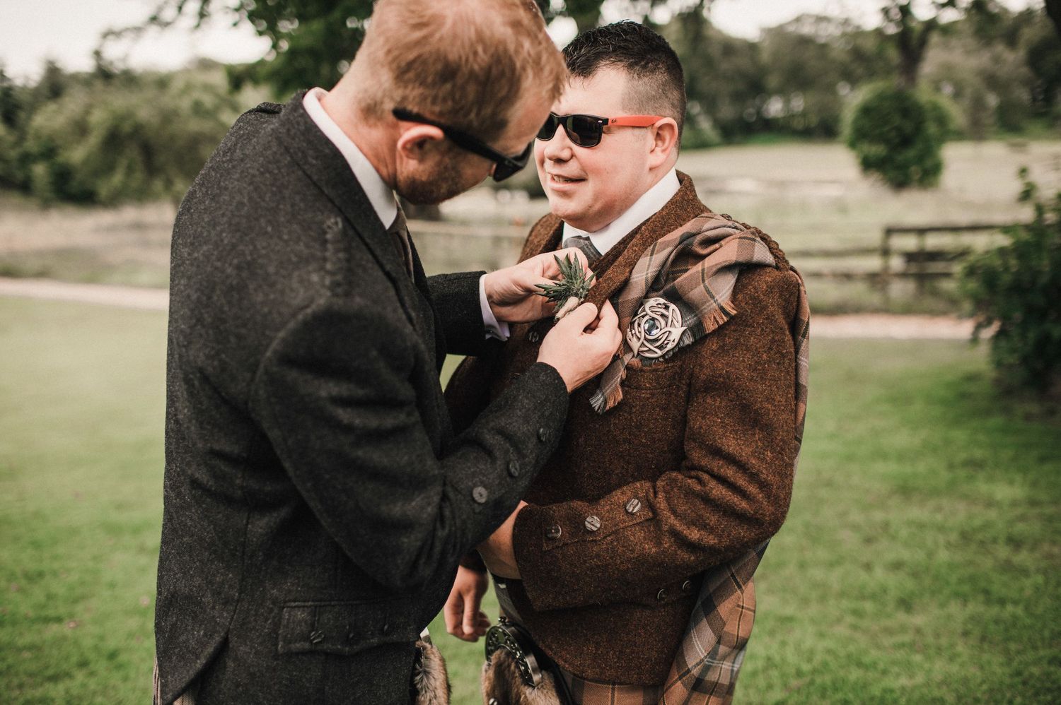 Two people in formal attire help adjust each other's boutonnieres outdoors.