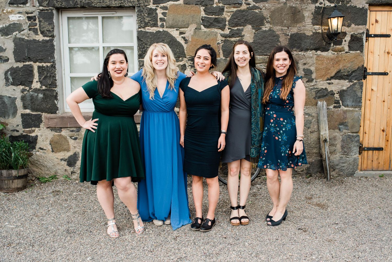 Group of wedding guests in formal dresses pose against stone wall exterior.