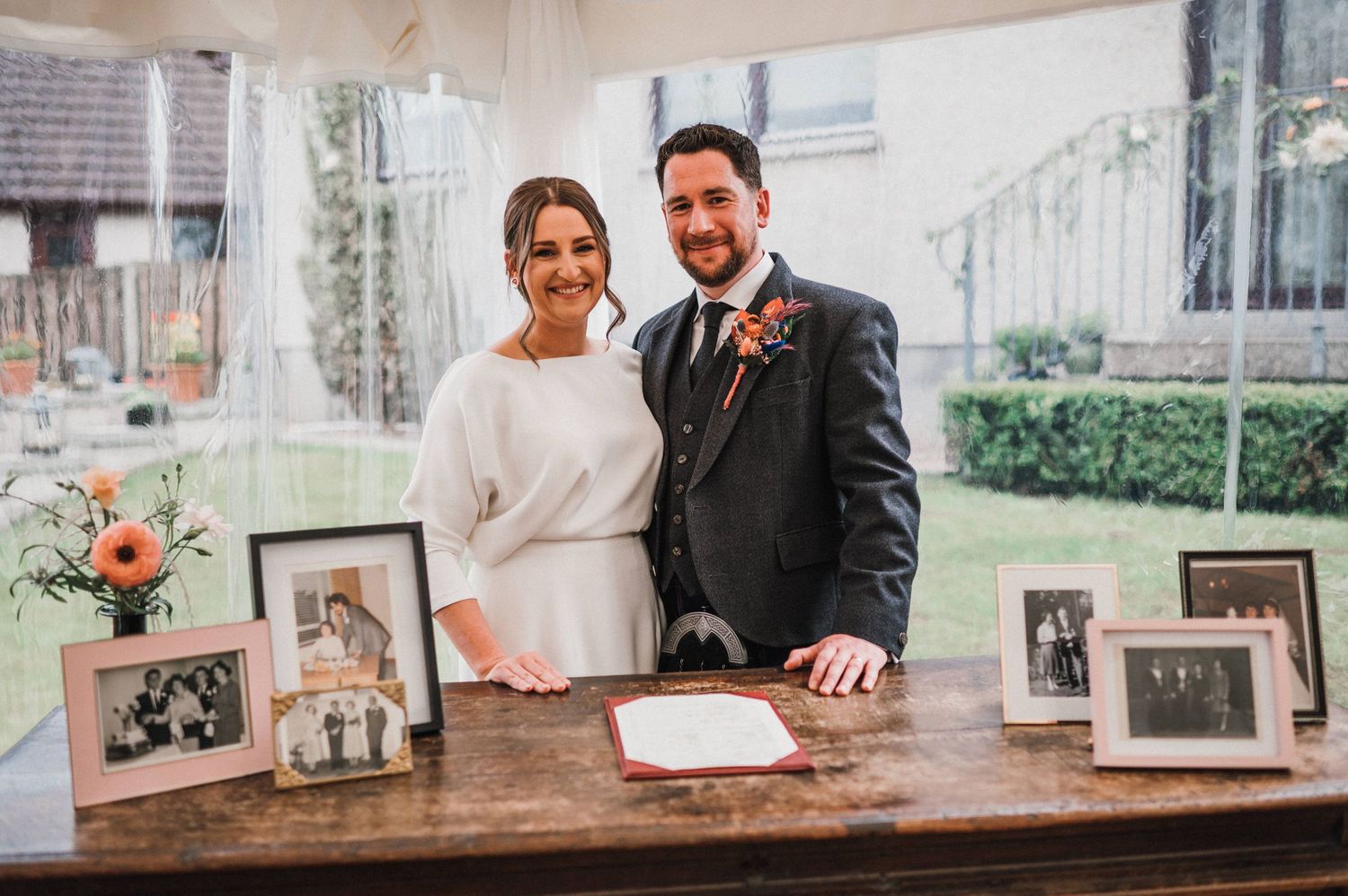Newlyweds pose behind antique table with family photographs and orange floral arrangements.