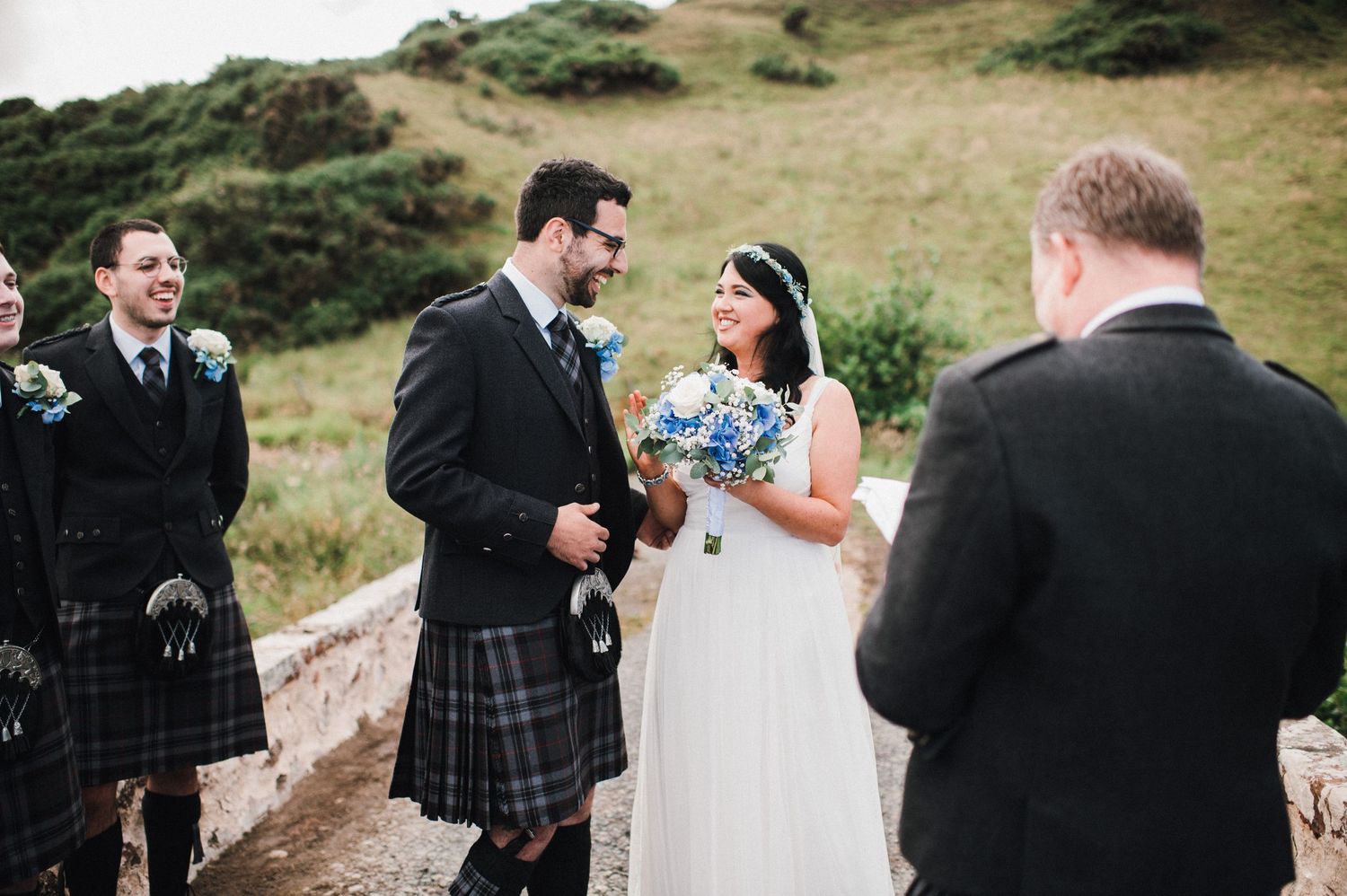 A traditional Scottish wedding ceremony with the groom in a kilt and bride in white on a scenic hillside path.