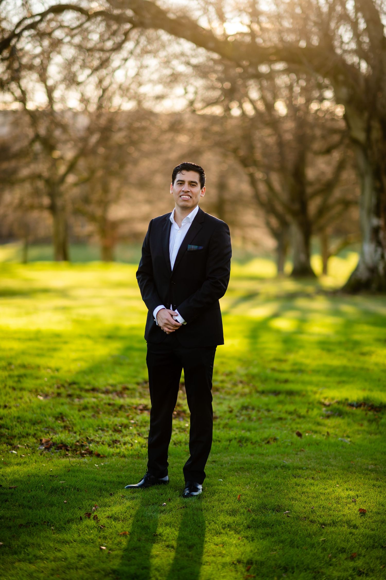 Person in formal attire standing on grass with trees and sunset lighting in background.