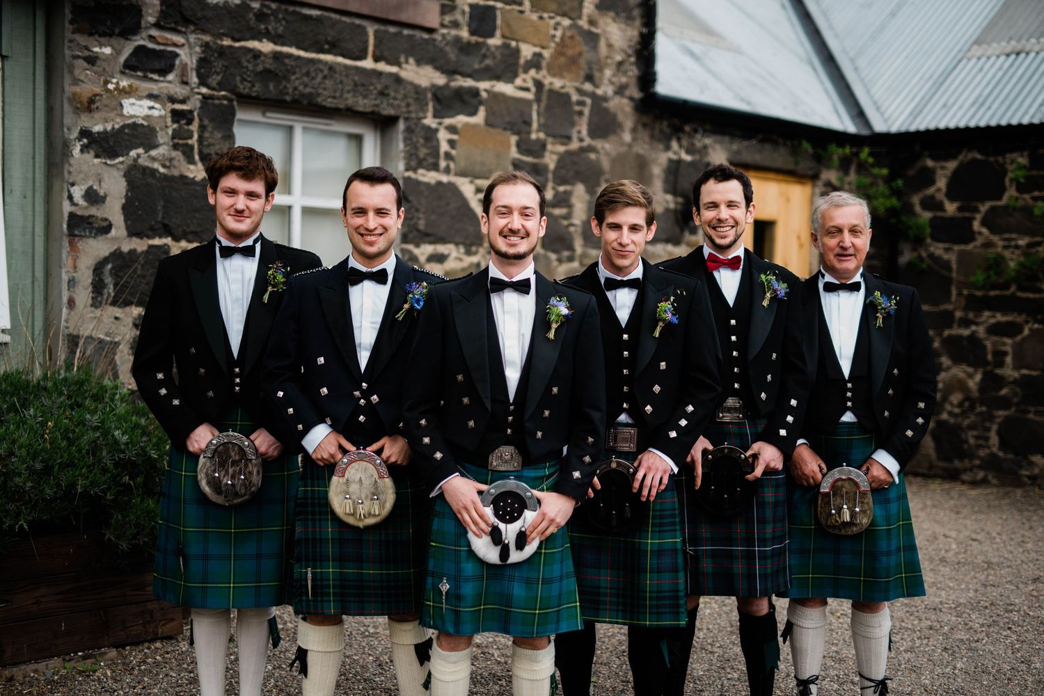 Groomsmen in traditional Scottish kilts pose together against stone wall backdrop.