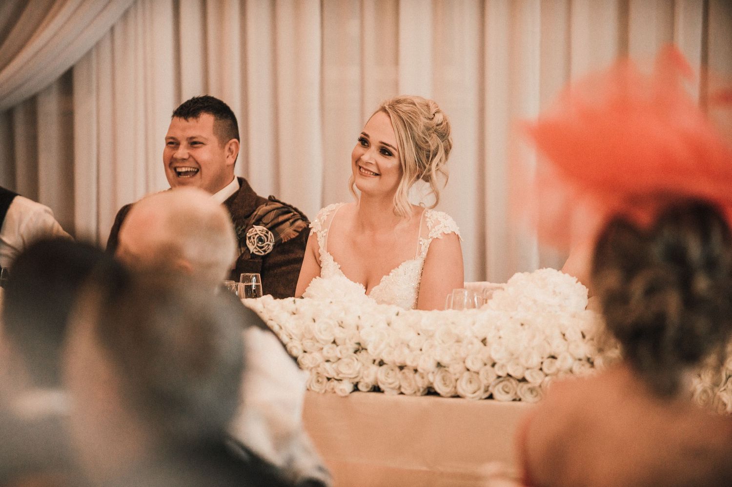 Wedding reception scene with guests seated at a decorated head table with floral arrangements.