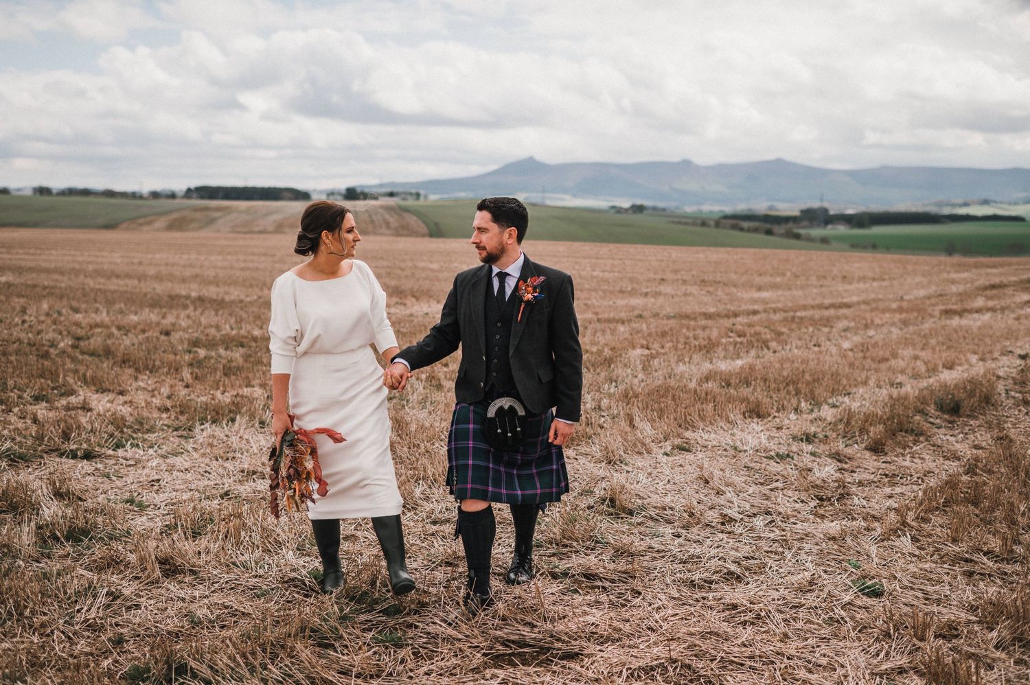 A couple in wedding attire walks through a rural field with the groom wearing a traditional Scottish kilt.