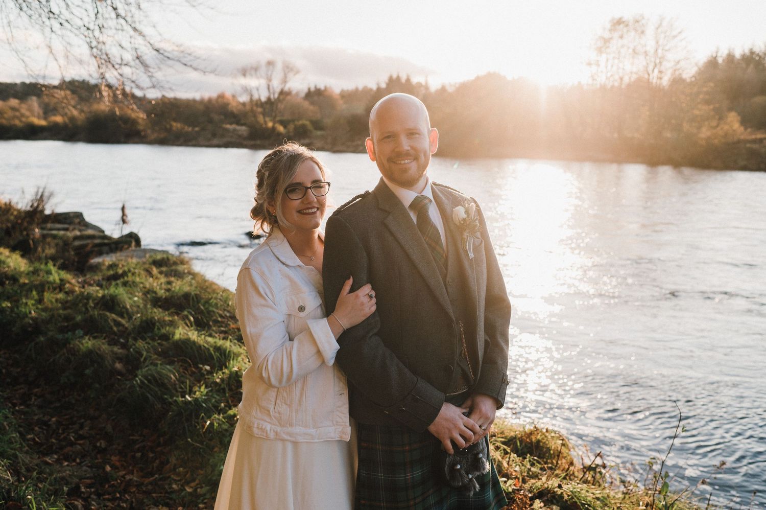 A wedding couple poses by a river during sunset wearing traditional Scottish attire.