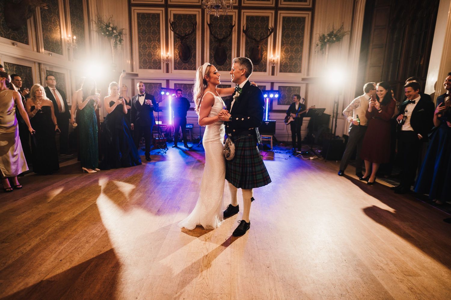 Couple dancing romantically on a wooden dance floor with blue lighting and onlookers at evening reception.