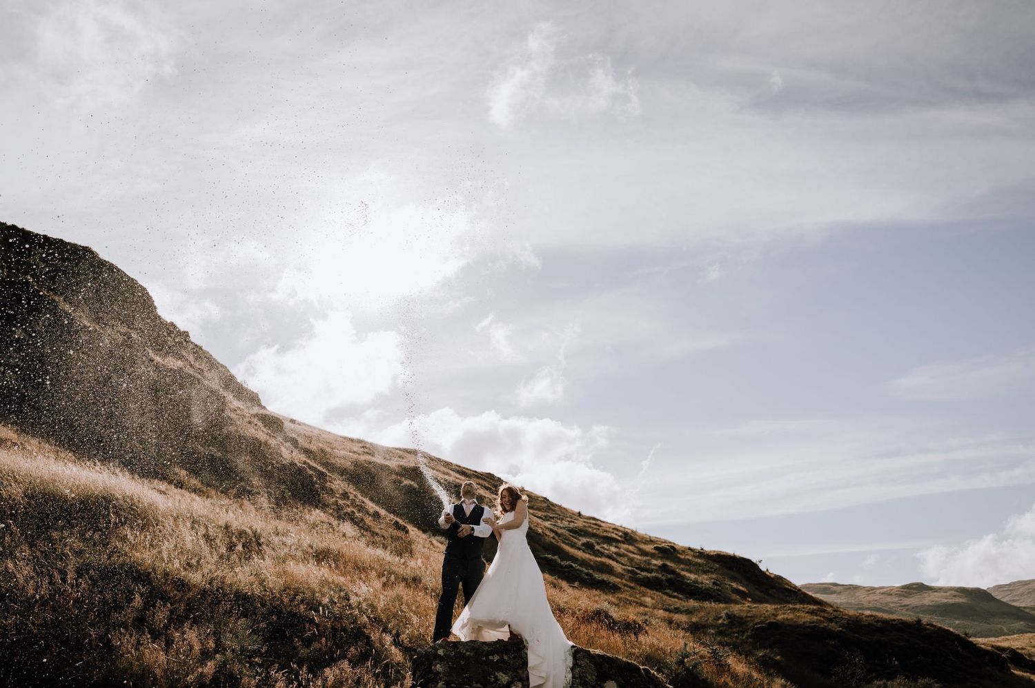 A wedding couple stands silhouetted against dramatic mountains and cloudy skies in a romantic outdoor setting.
