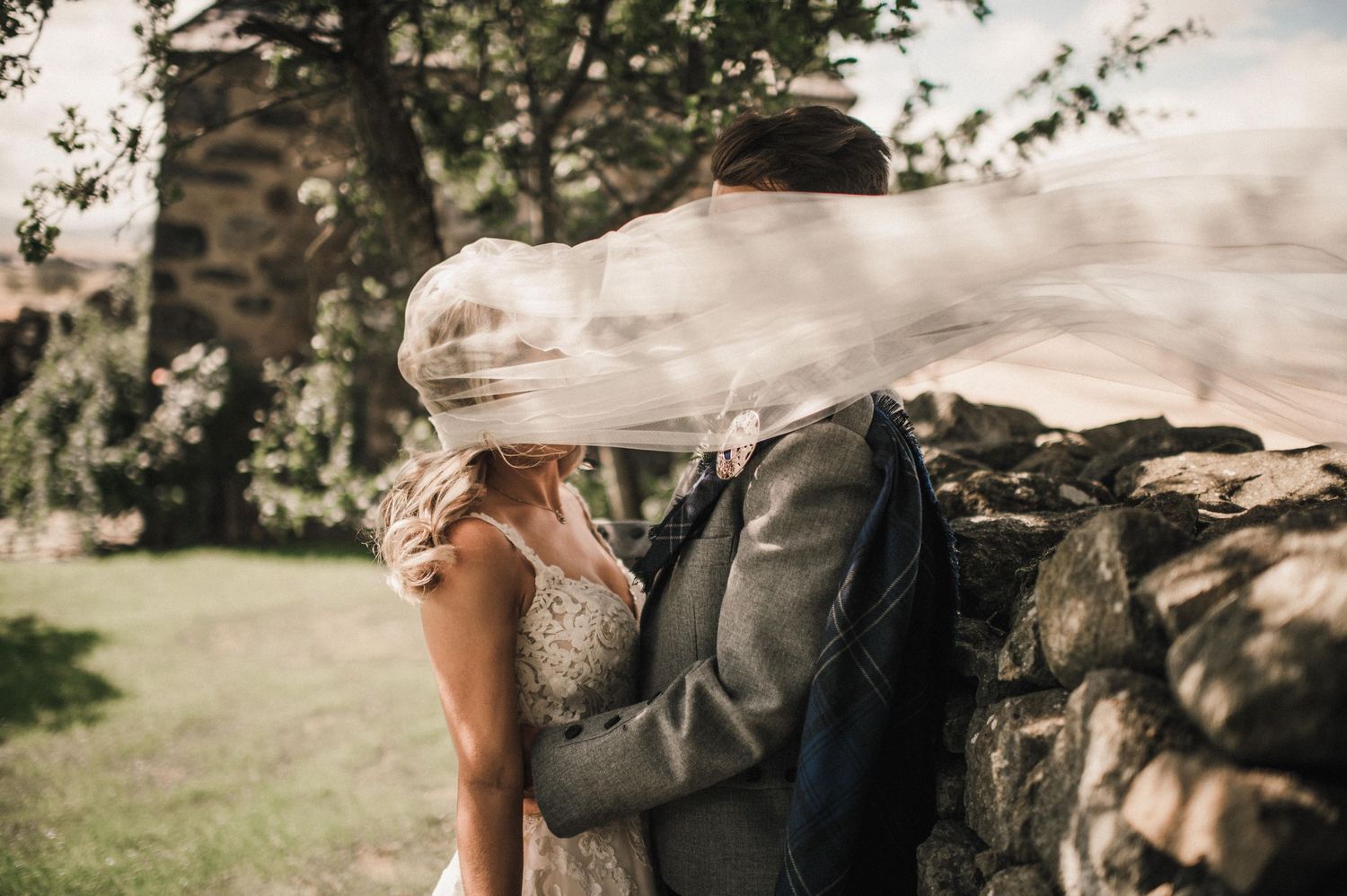Wedding veil flows in the wind as couple embraces by stone wall.
