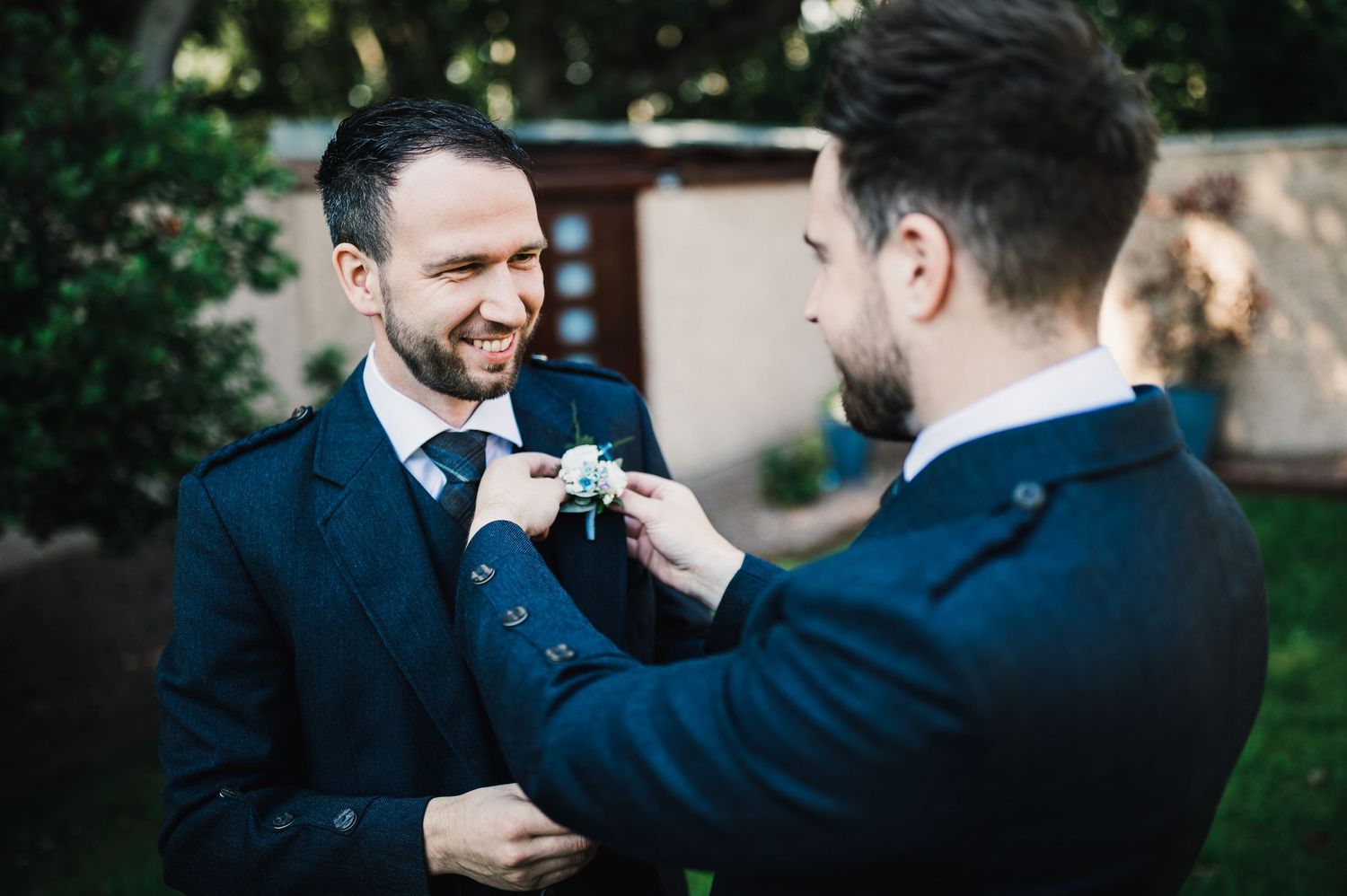 Two men in navy suits adjust each other's buttonhole flowers outdoors.