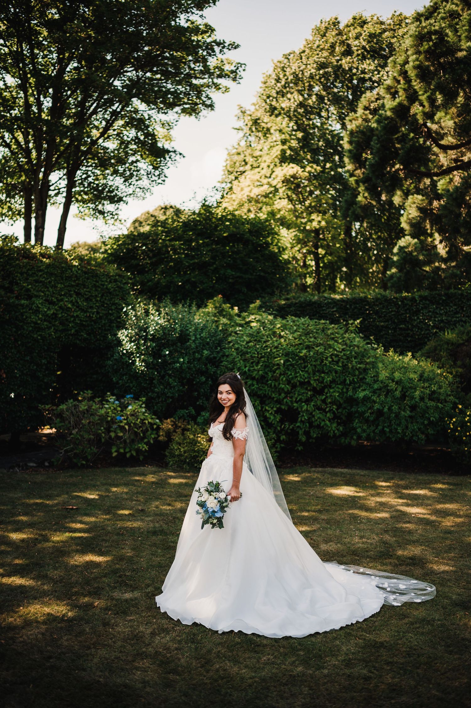 A bride in a flowing white gown and veil poses gracefully in a garden setting with sunlit trees in the background.