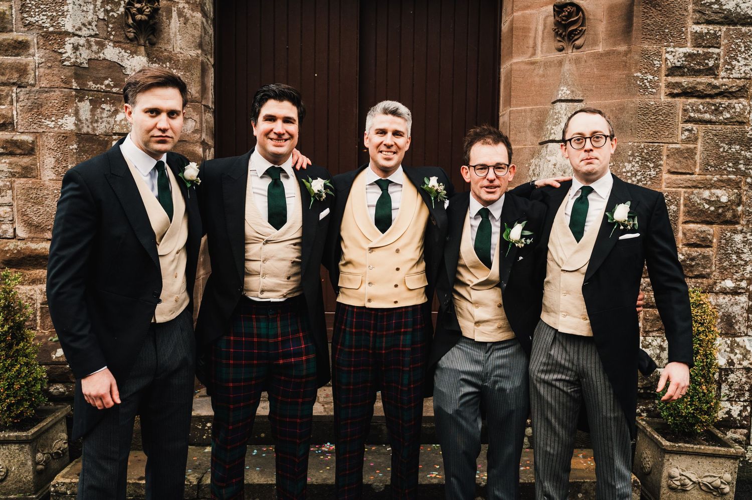 Groomsmen in matching kilts and waistcoats pose together at church entrance.