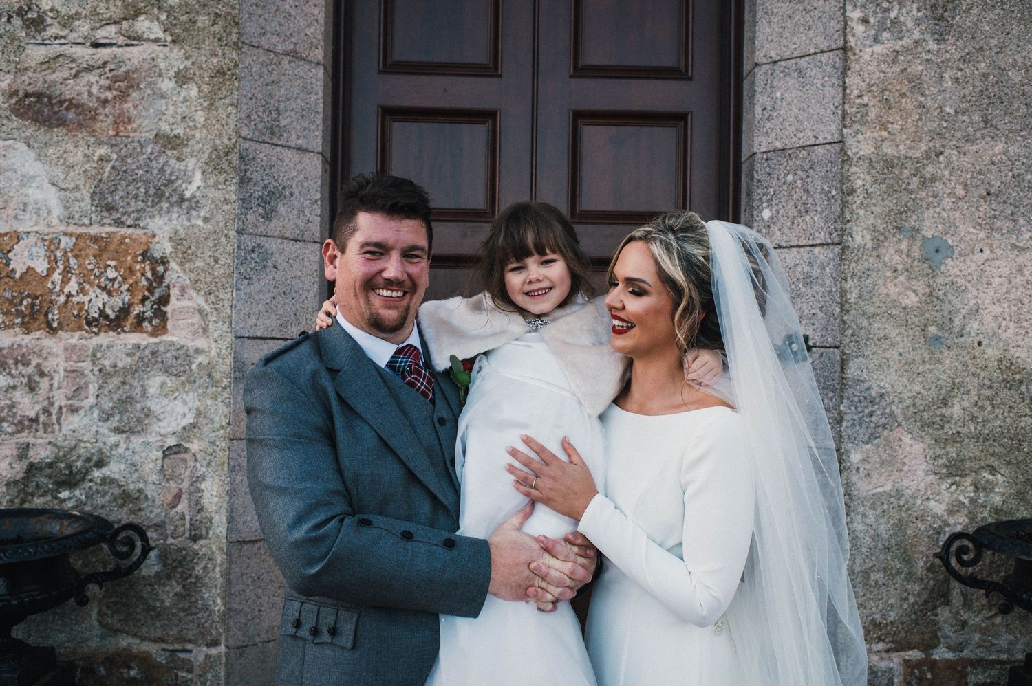 Wedding couple with young flower girl at stone church entrance.