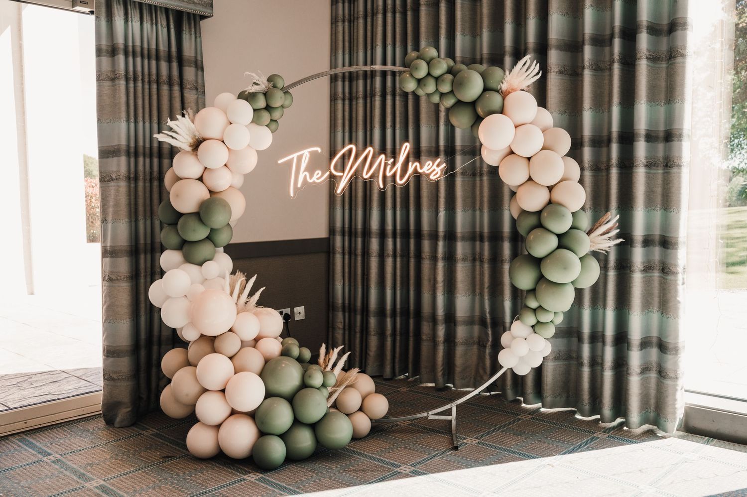 A decorative arch with pink and sage balloons frames a neon wedding sign.