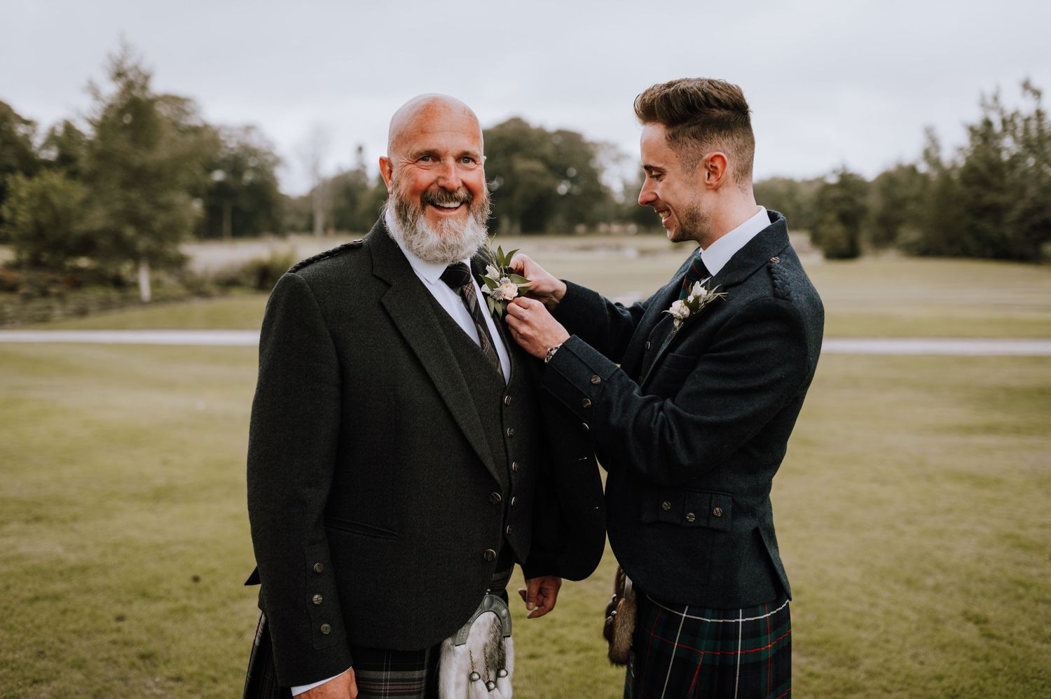 Young man in Scottish attire adjusting boutonniere on older gentleman's lapel.