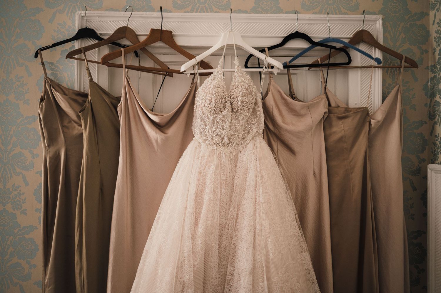 A white wedding gown hangs among neutral bridesmaid dresses on a clothing rack.