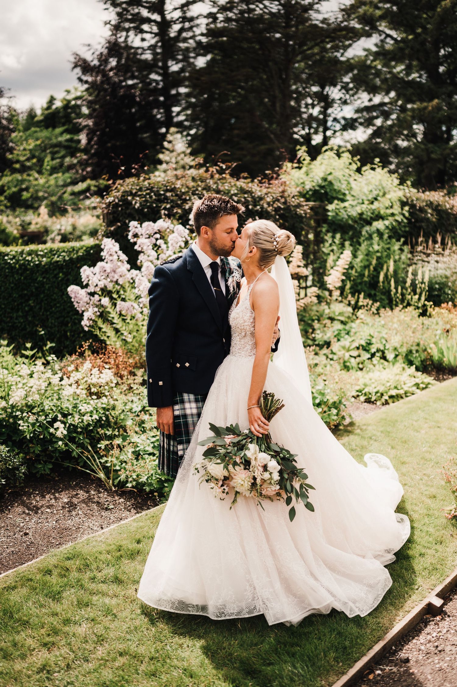A scenic garden wedding portrait featuring Scottish formal wear and an elegant white bridal gown.