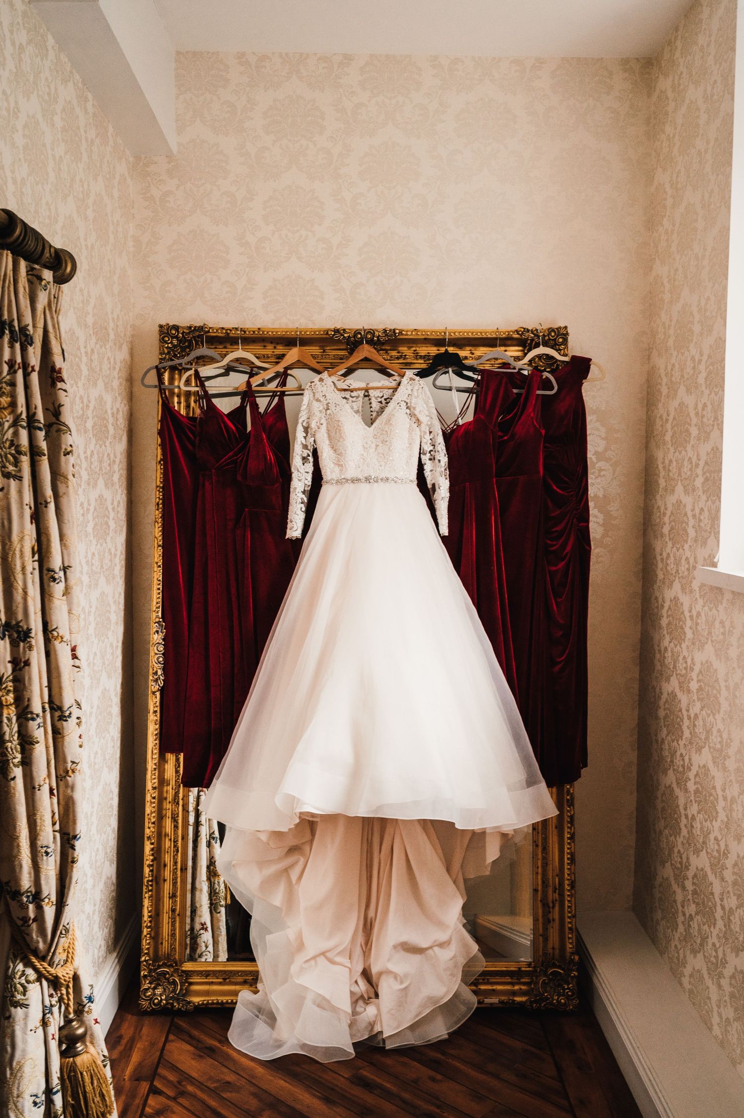White lace wedding gown hanging on gold frame mirror with burgundy bridesmaid dresses.