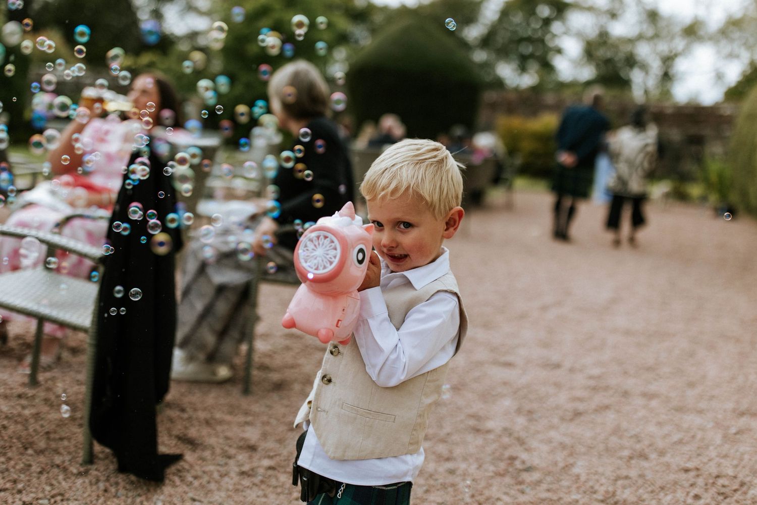 A young child in a vest blows bubbles at an outdoor event.