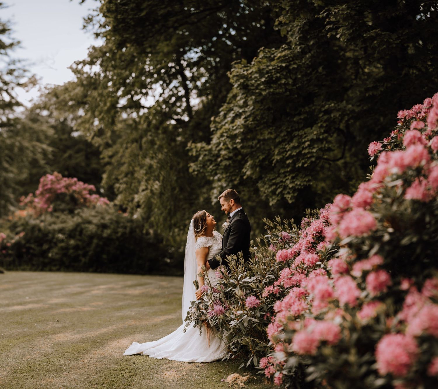 Wedding couple poses beside blooming pink flowers in a lush garden setting.
