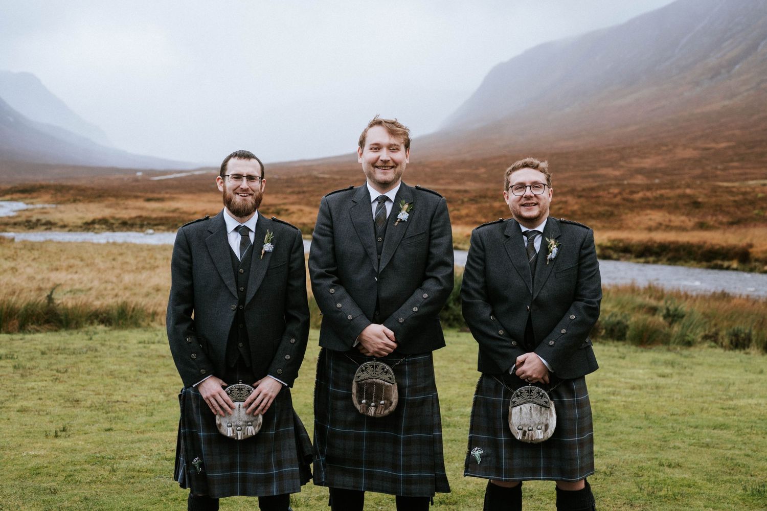 Three men in traditional Scottish kilts and formal wear standing in a misty highland landscape.