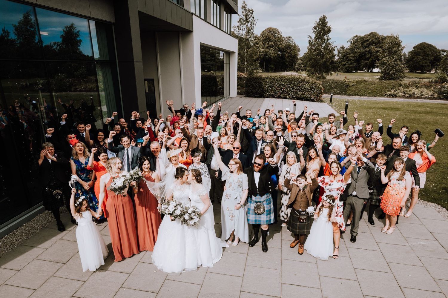 Large wedding group photo of guests and wedding party gathered outside a modern building.