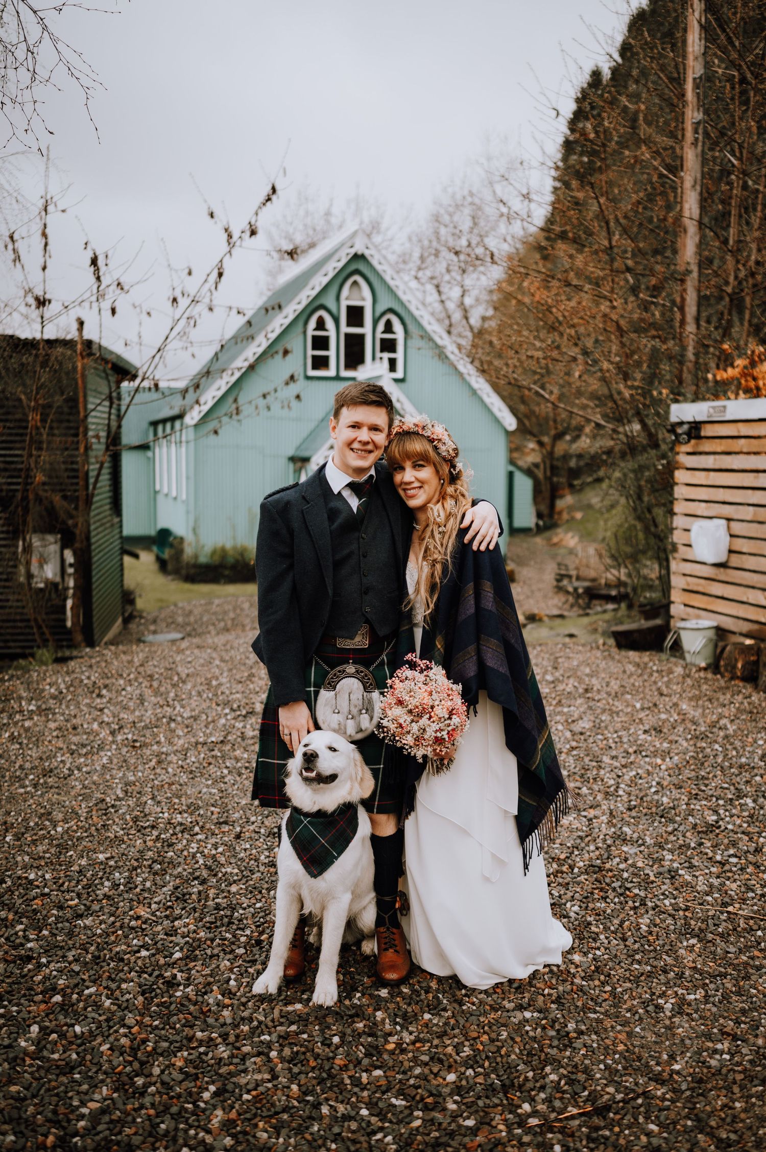 White dog wearing a black bandana poses with wedding couple in front of a mint green chapel building on an autumn day.