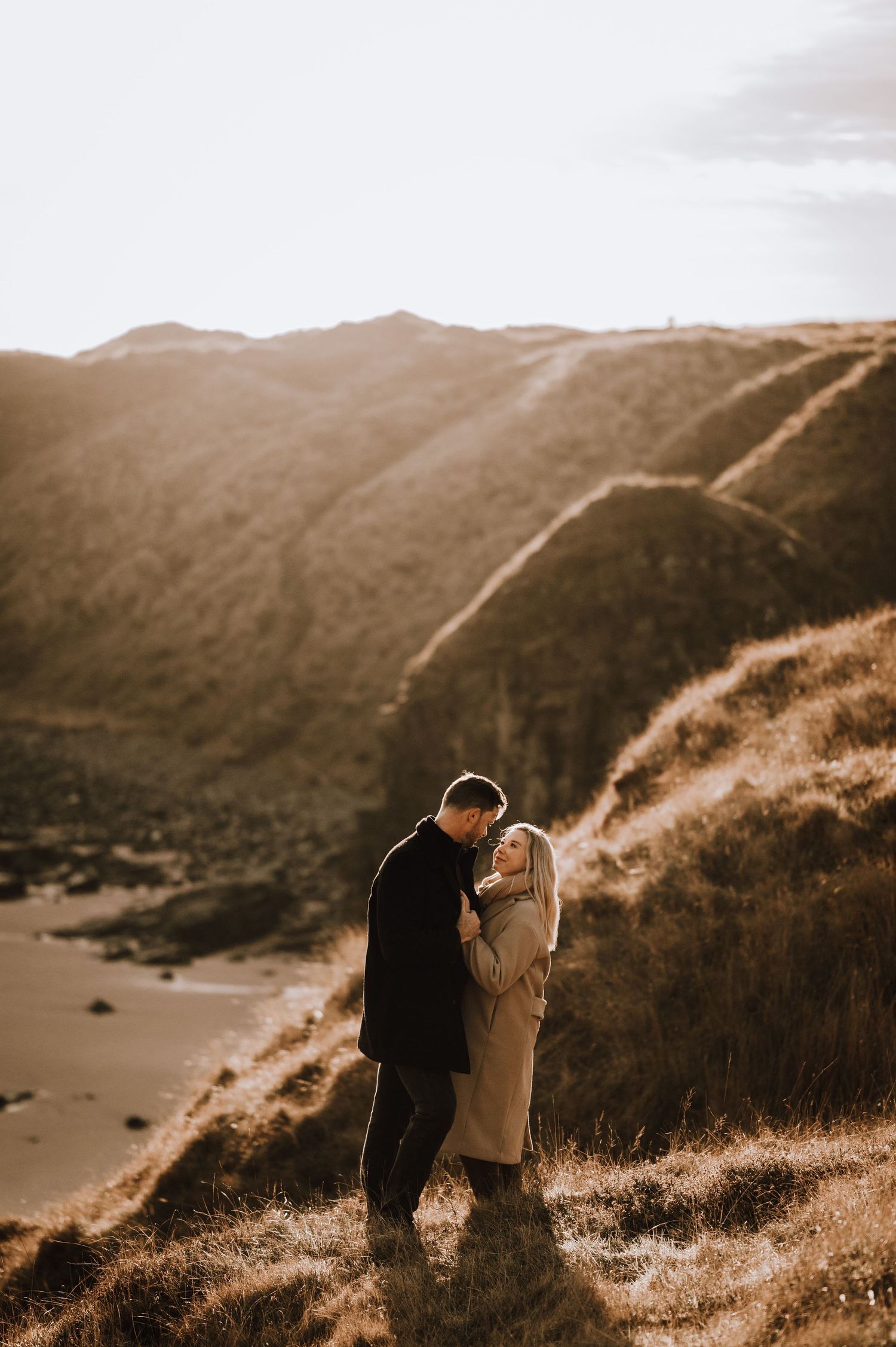 Two people stand close together on a hillside path overlooking a scenic coastal view.