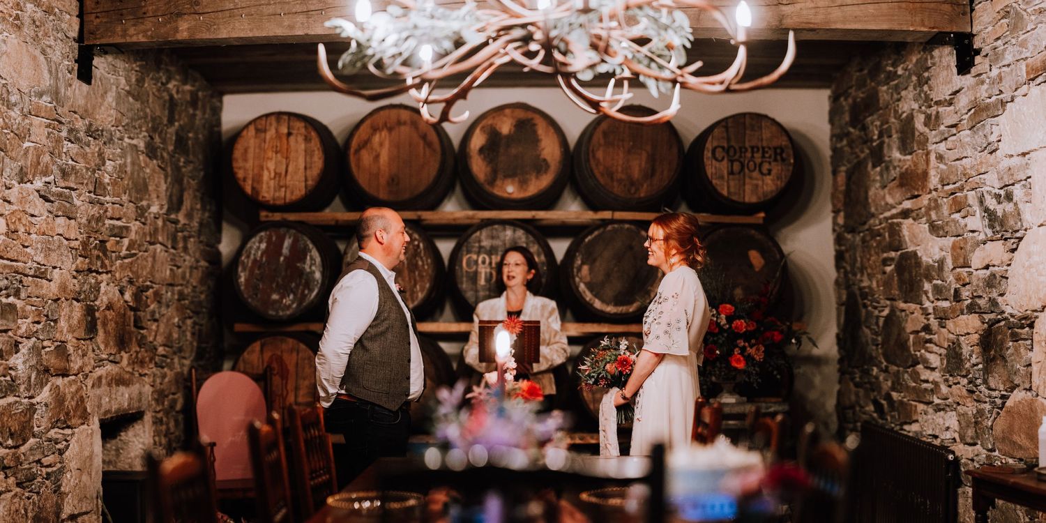 Rustic wine cellar wedding ceremony setup with wooden barrels and chandelier.
