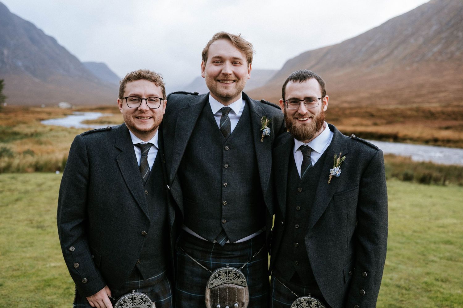 Groomsmen in traditional Scottish kilts and formal wear stand together against a mountainous Highland backdrop.