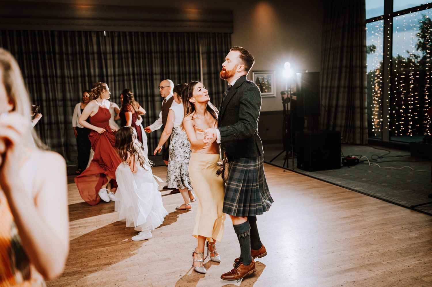 Wedding guests dance at a reception, with a person in traditional Scottish kilt visible.