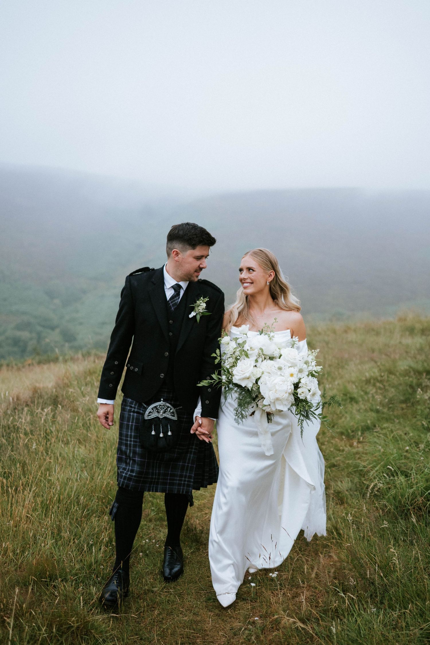 Couple walks through misty Scottish highlands in traditional wedding attire.