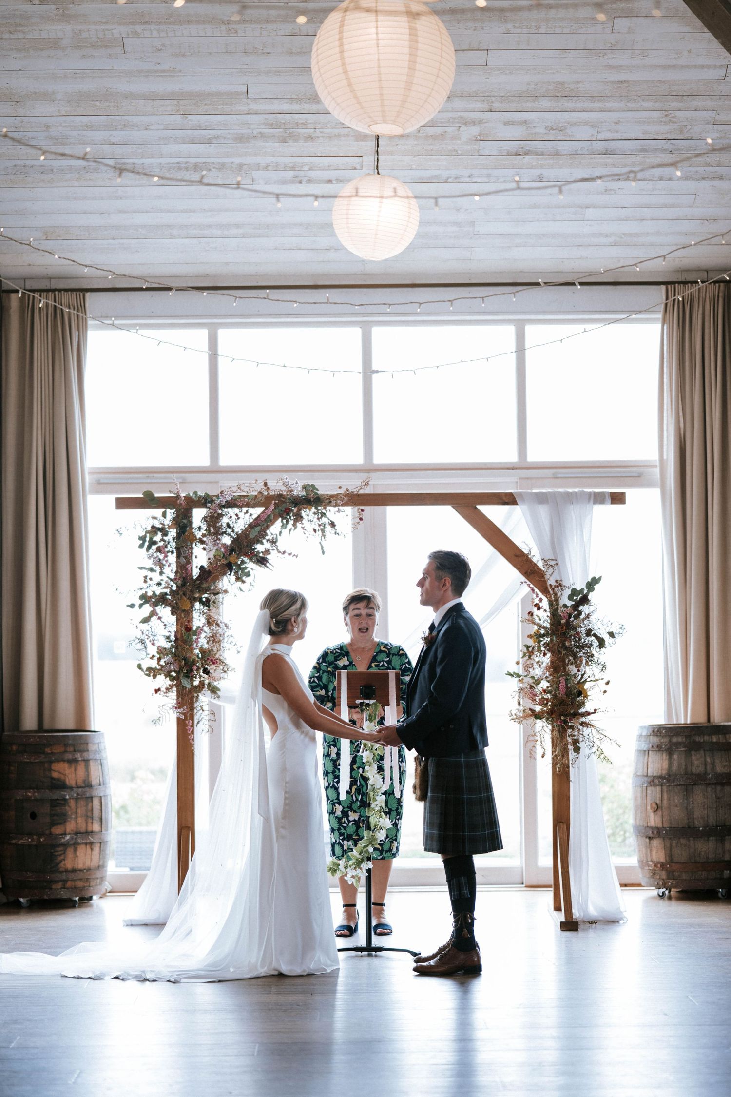 Wedding ceremony taking place under wooden arch with paper lanterns and barrel decor in bright indoor venue.