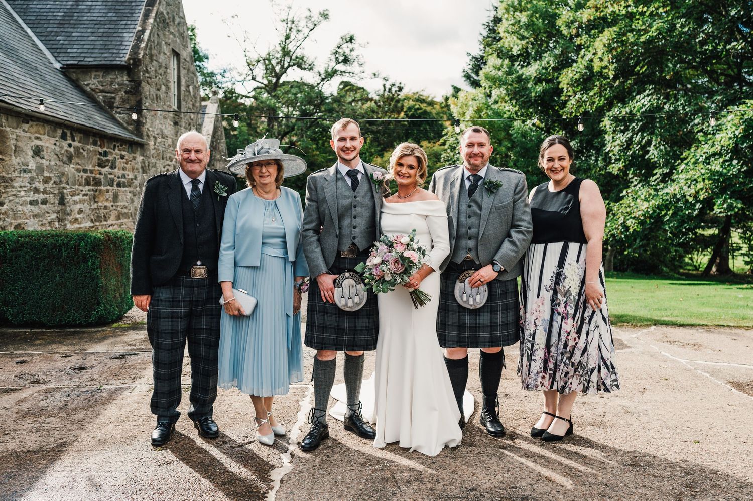 Wedding party posing outside historic stone building with greenery in background.