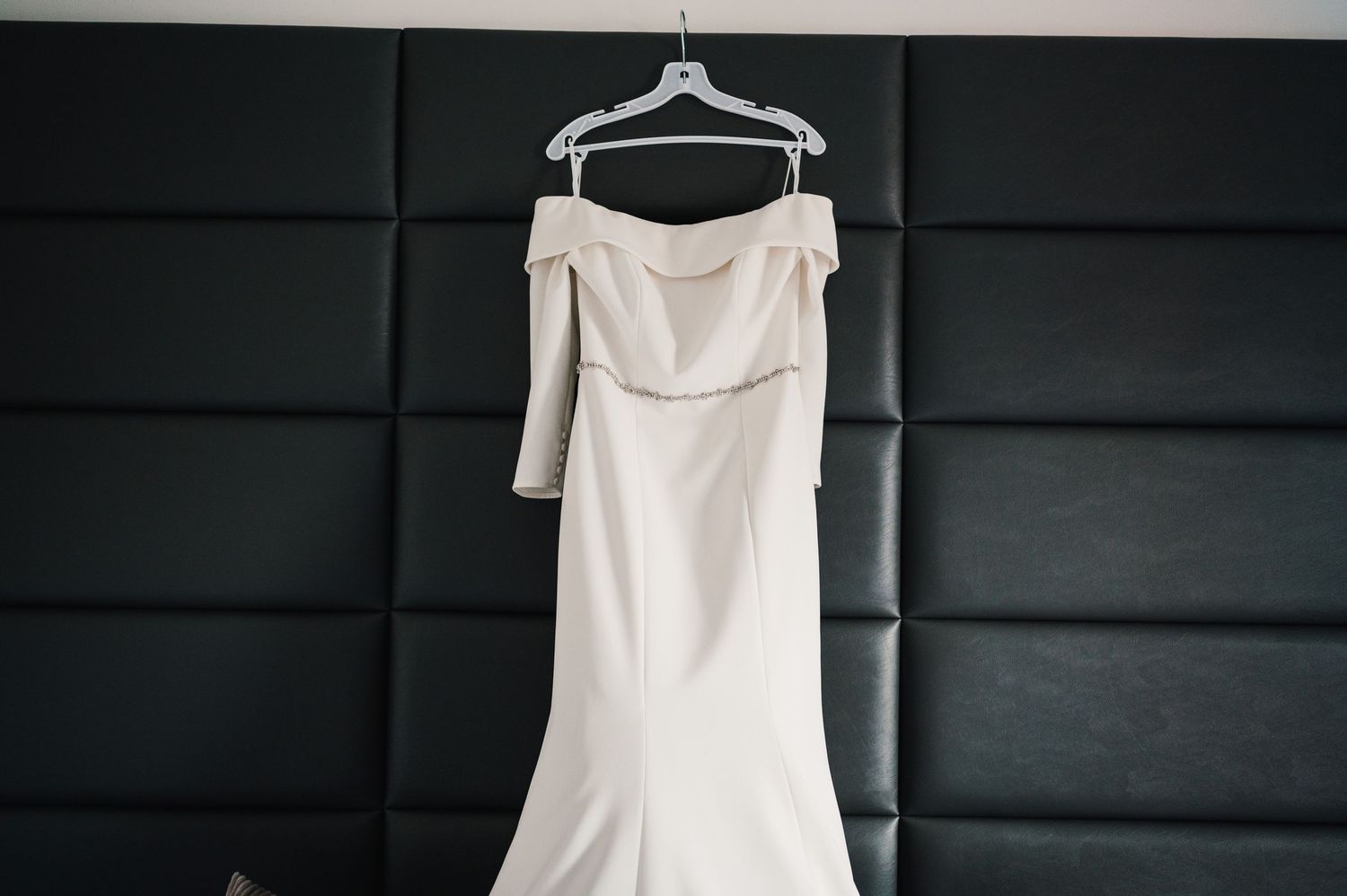 An elegant white off-shoulder wedding dress with long sleeves hangs on a hanger against a dark wall.